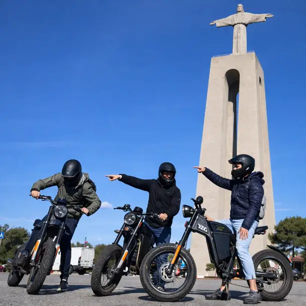 Three riders on electric motorcycles pointing toward the Cristo Rei monument under a clear sky during a Lisbon bike tour.