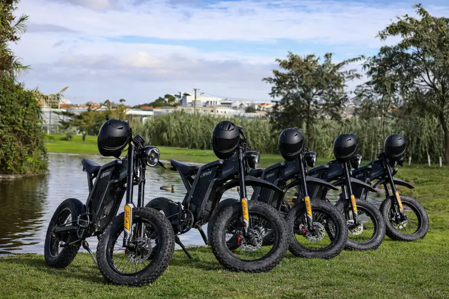 Five black electric dirt bikes lined up on grass by the water, showcasing off-road power during a Lisbon bike tour.